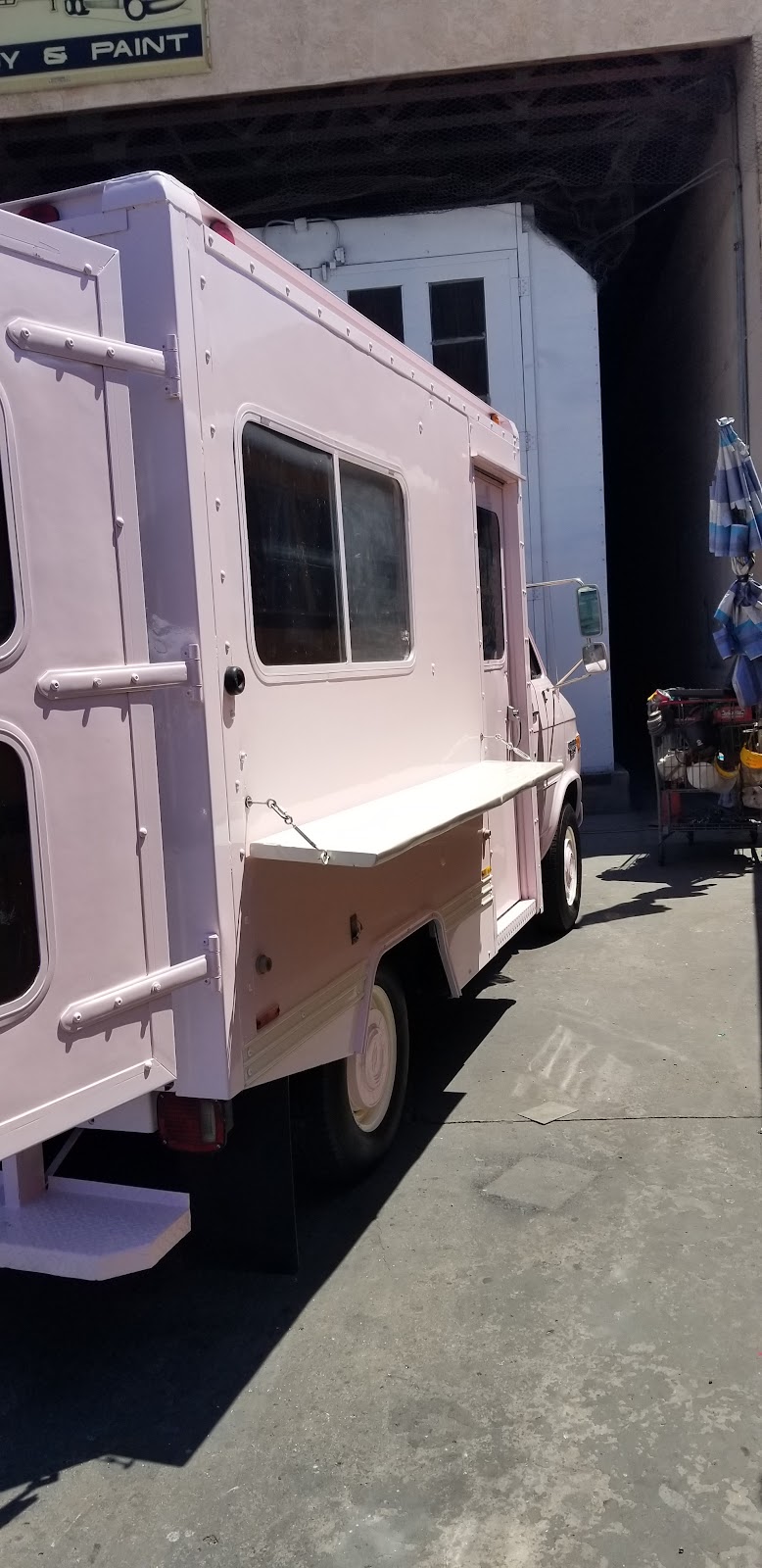 Pastel pink box-body food and concession truck with a fold-down serving counter, parked at the shop's bay door under the 'Body & Paint' sign